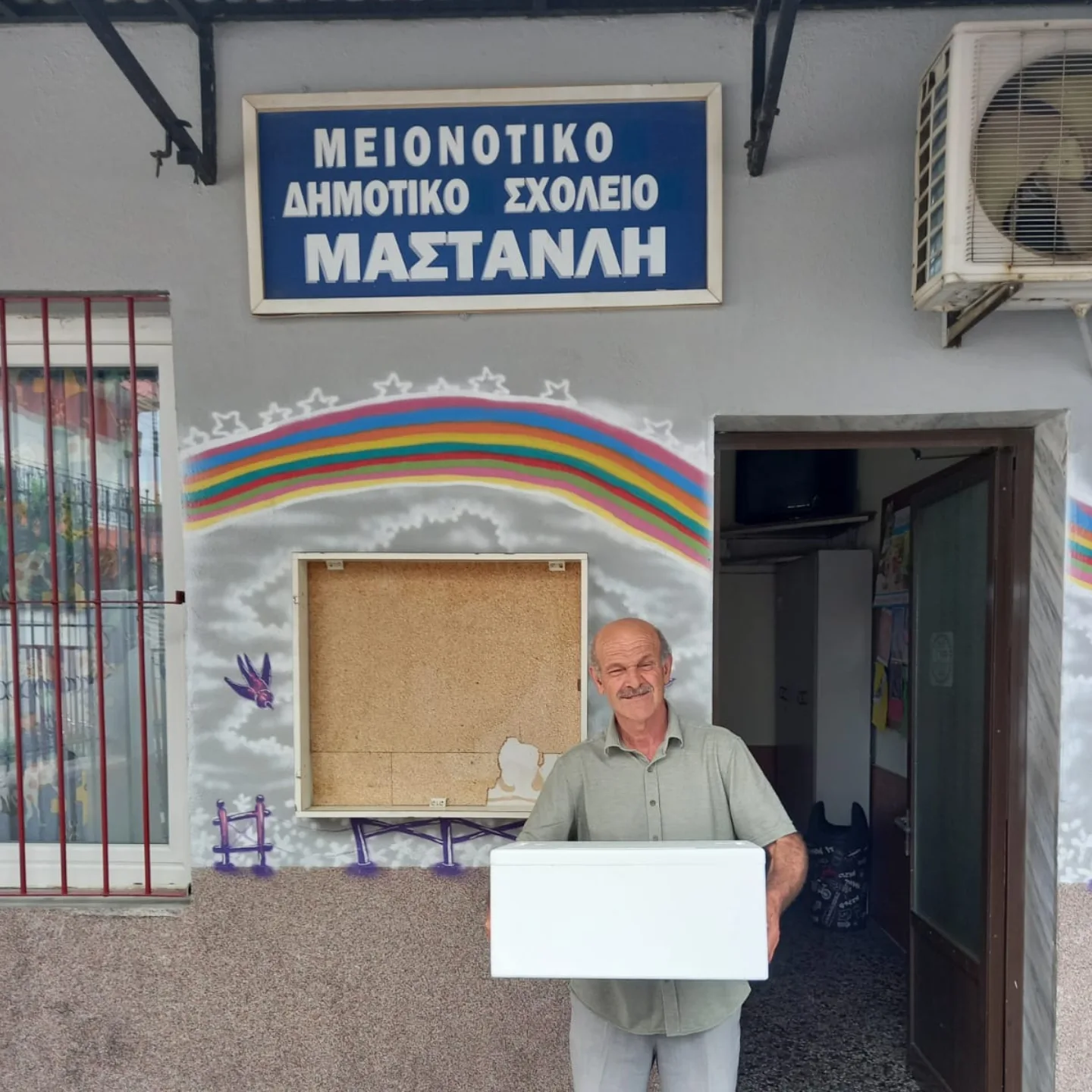 Ενήλικας κρατά λευκό φορητό ψυγείο μπροστά από την πινακίδα του Μειονοτικού Δημοτικού Σχολείου Μαστανλή.